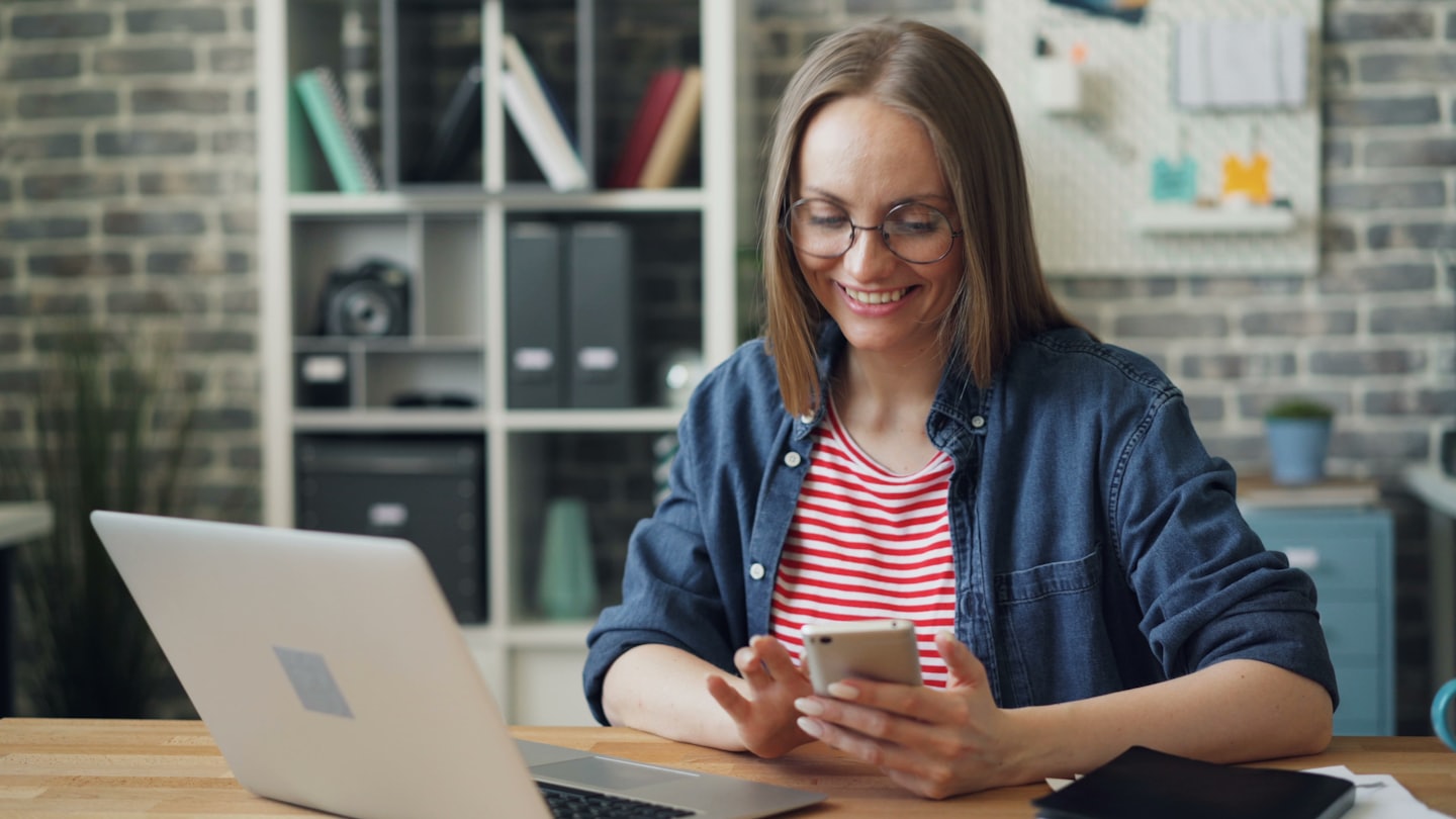 Woman smiling while using her smartphone at a home office desk.